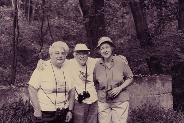 Dorothy Miller, wearing binoculars around her neck and posing for a photo while birding with 2 companions in the forest.