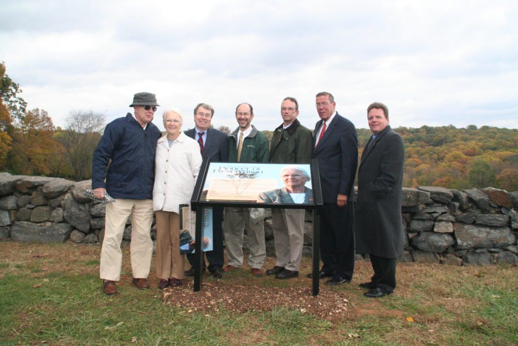 Lynn Williams, smiling, and surrounded by members of the parks and recreation community at the dedication of an educational wayside  display highlighting to her life’s work. Standing atop a hill which overlooks an expanse of woodland. Iconic rock walls of Brandywine Creek State Park pictured in background.