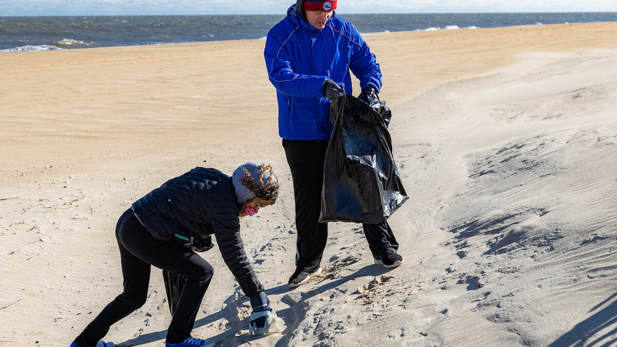 Two people gather trash on an empty beach in the winter. A man in a blue coat, right, holds a trash bag while a woman in a black coat, left, bends to pick up litter from the dunes. The Atlantic Ocean is calm with small waves in the background.