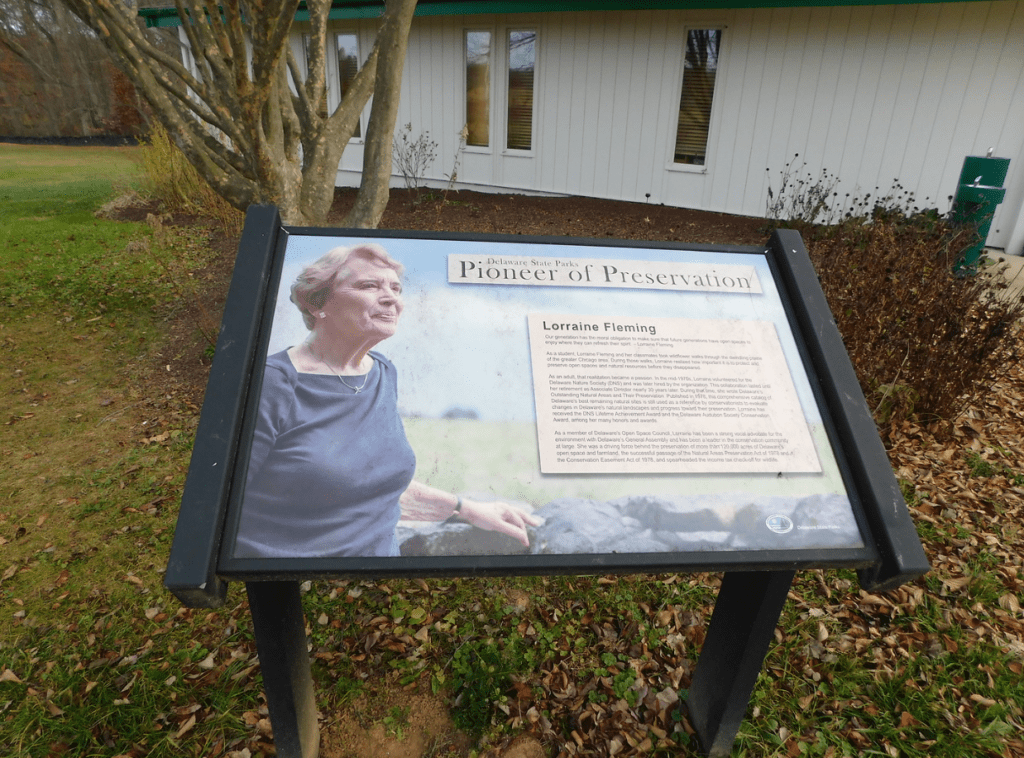 The historical marker dedicated to Lorraine Fleming at Brandywine Creek State Park's Nature Center, in honor of her environmental activism. The marker depicts Lorraine standing next to the iconic stone walls of the park and includes a text box describing her contributions to the environmental activism. The nature center is in the background, and the ground is littered with early fall leaves.