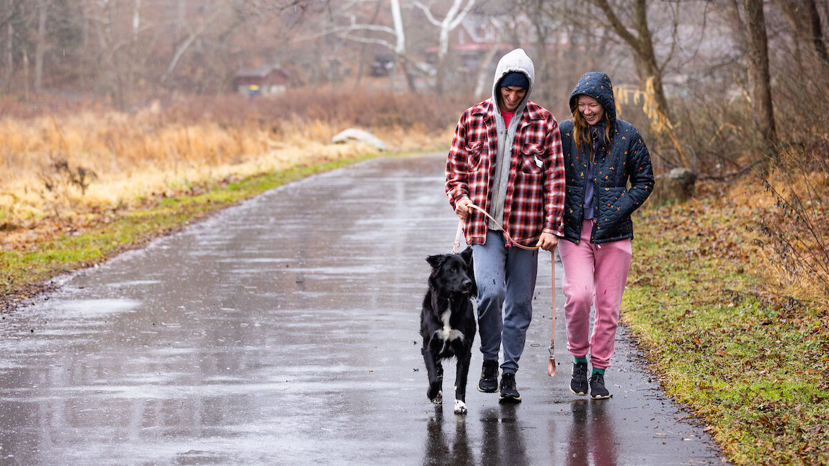 Two park users hike on a paved trail on a rainy winter day, laughing to themselves. Their black and white mid-sized herding dog is walking on a leash in the center of the photo, alert to movement off of the trail to the right.