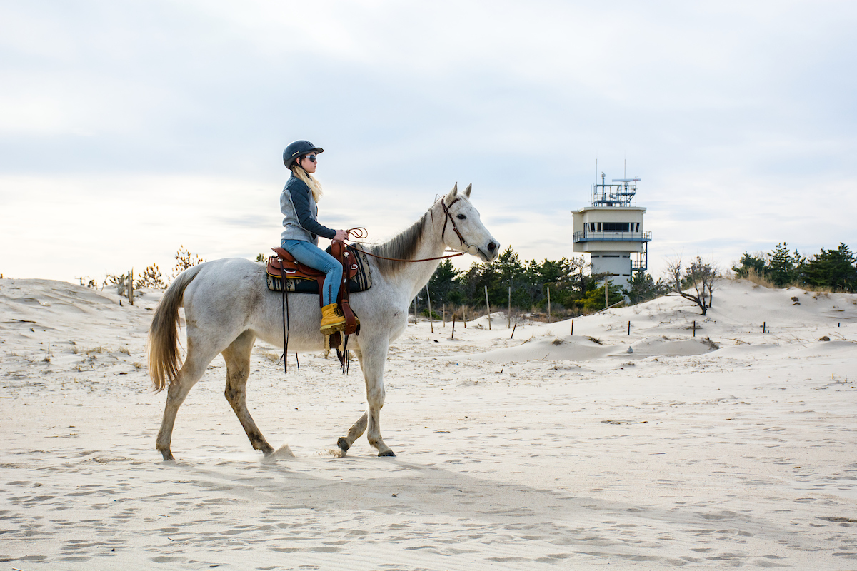 Young woman on a white horse rides atop the sand at Cape Henlopen State Park's Point. In the foreground, the horse and rider are parallel to the camera and face the right of the image looking into the distance. In the background, rolling sand dunes and a tall, concrete structure - the pilot station - are visible. The image is taken in the early morning light, softening the edges of the scene.