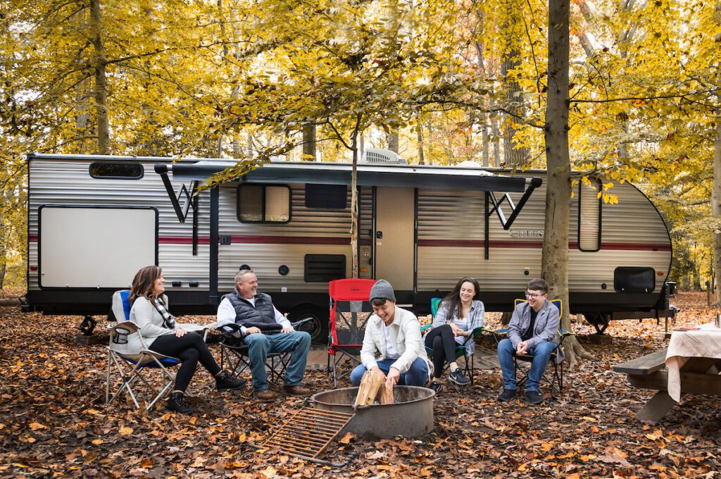 A family of 5 in fall attire are sitting in a row of camping chairs in front of a fire with their RV parked behind them. In the background are trees of yellow hues and scattered leaves of the forest floor.