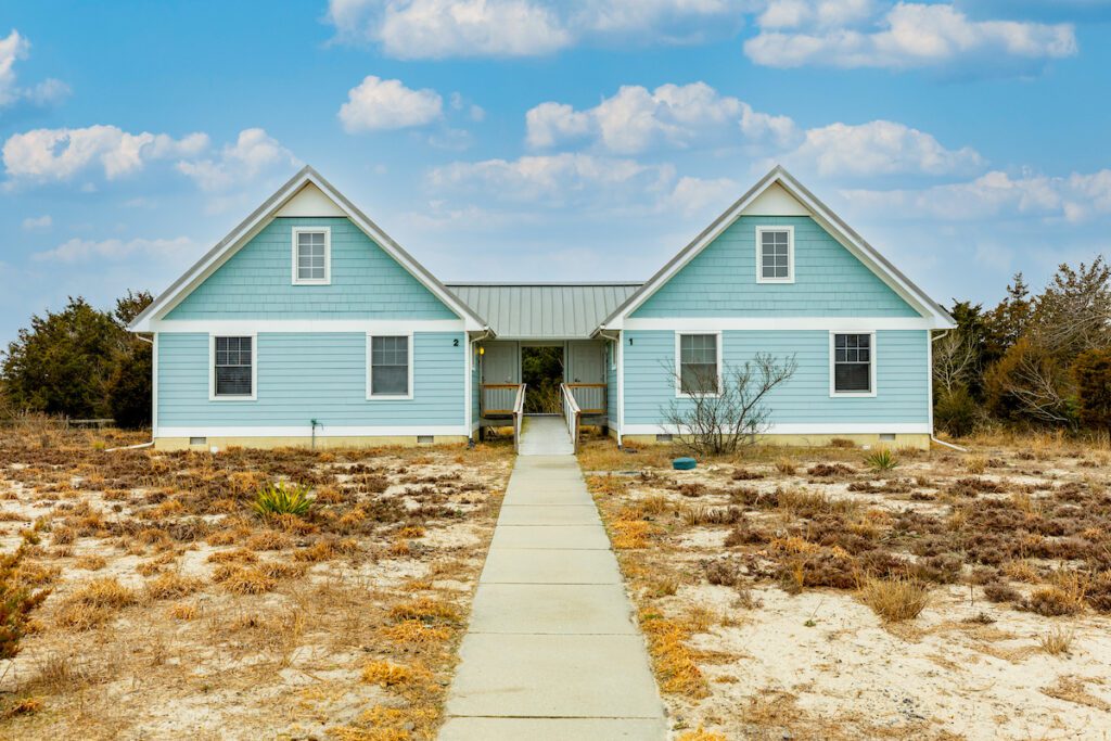 A light-blue symmetrical cottage tucked by the water's edge. The front of the cottage features a lightly sandy open space. 