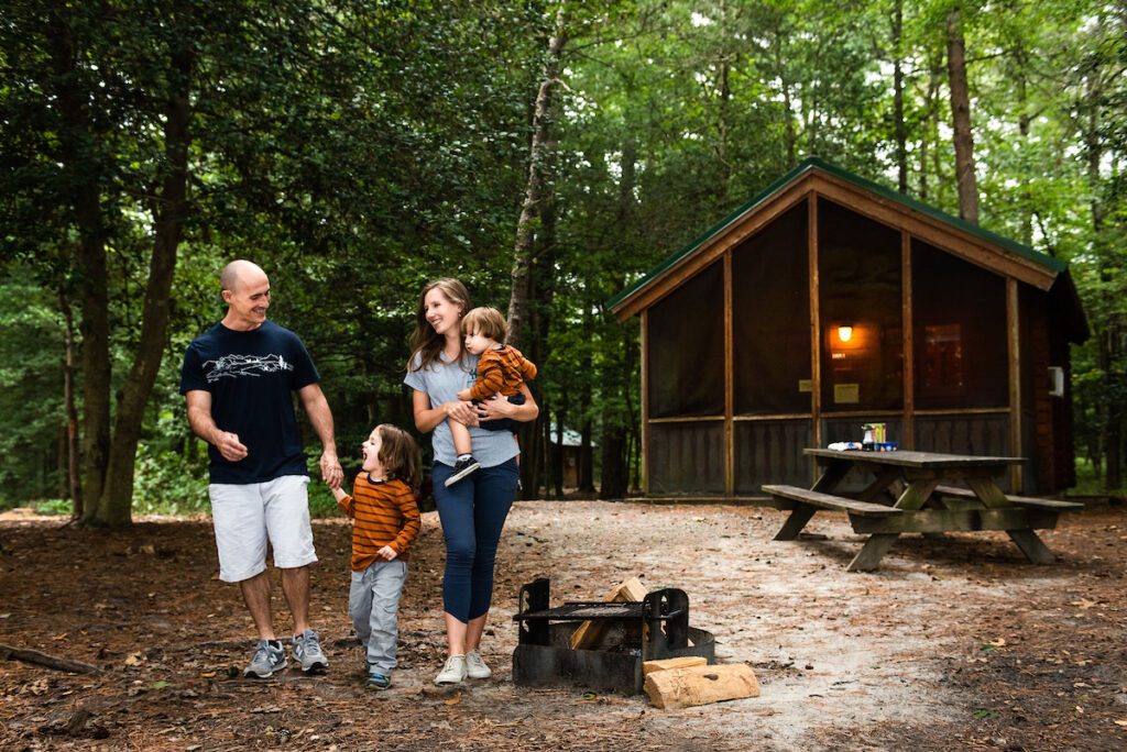 A smiling family of two adults and two children, walking outside their cabin in Trap Pond State Park with lush trees in the background.