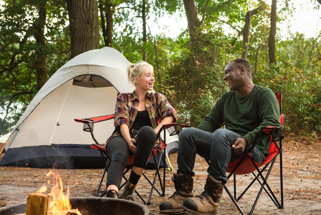 A couple in a forest are sitting in camping chairs and enjoying the fire with their camping set-up seen behind them.