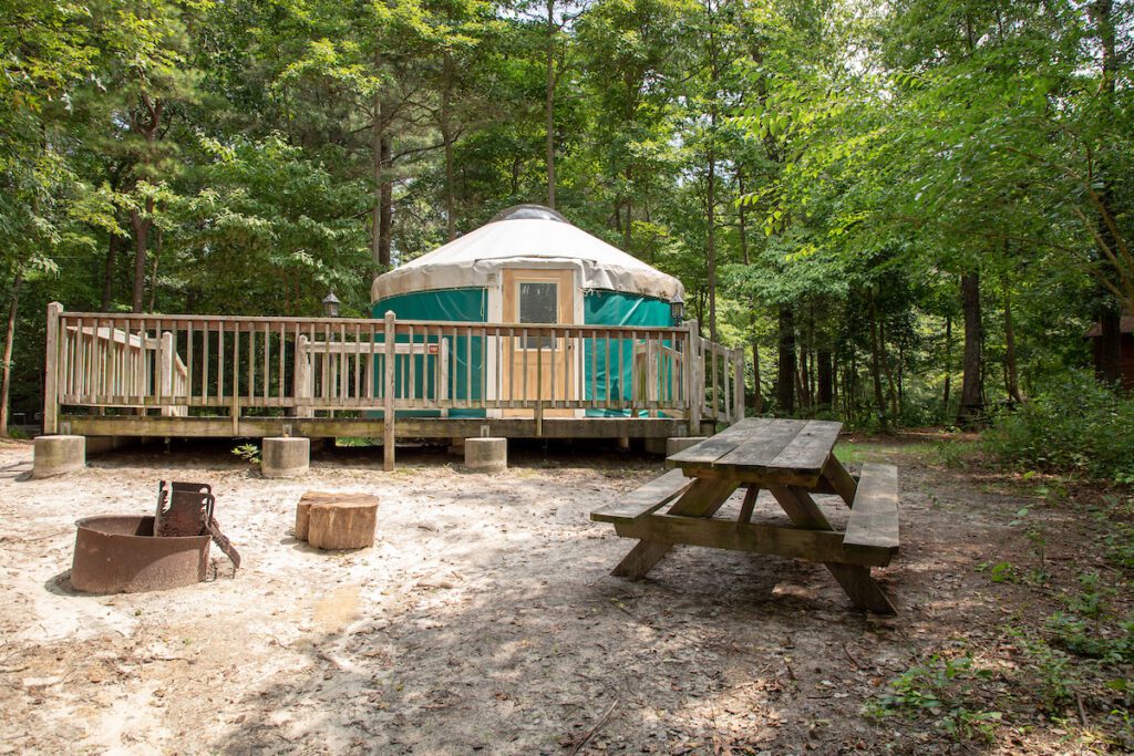 A dark green yurt is positioned on a wooden deck platform, outside features a fire pit and picnic table, surrounding the tent are tall green trees.