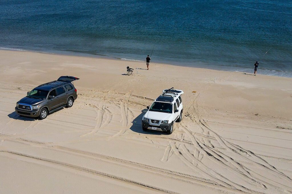 2 men surf fishing near the shore, behind them are 2 SUVs parked on the beach.