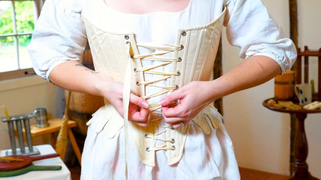 A woman lacing up a cream-colored stay over a white shift with puffy sleeves. The stay has a crisscross pattern, the woman is actively tightening the lacing.