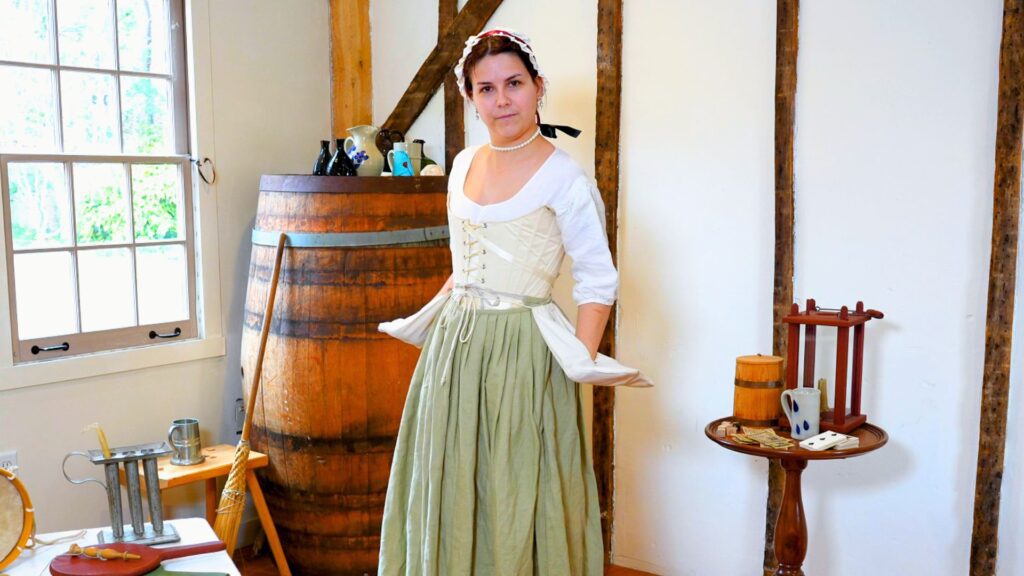 A woman dressed in historical attire standing in an old fashion interior setting with historical decor. She wears a white shift, white bonnet, cream stay and olive petticoat. She stands with her hands in tie-on pockets and she continues to layer her outfit.